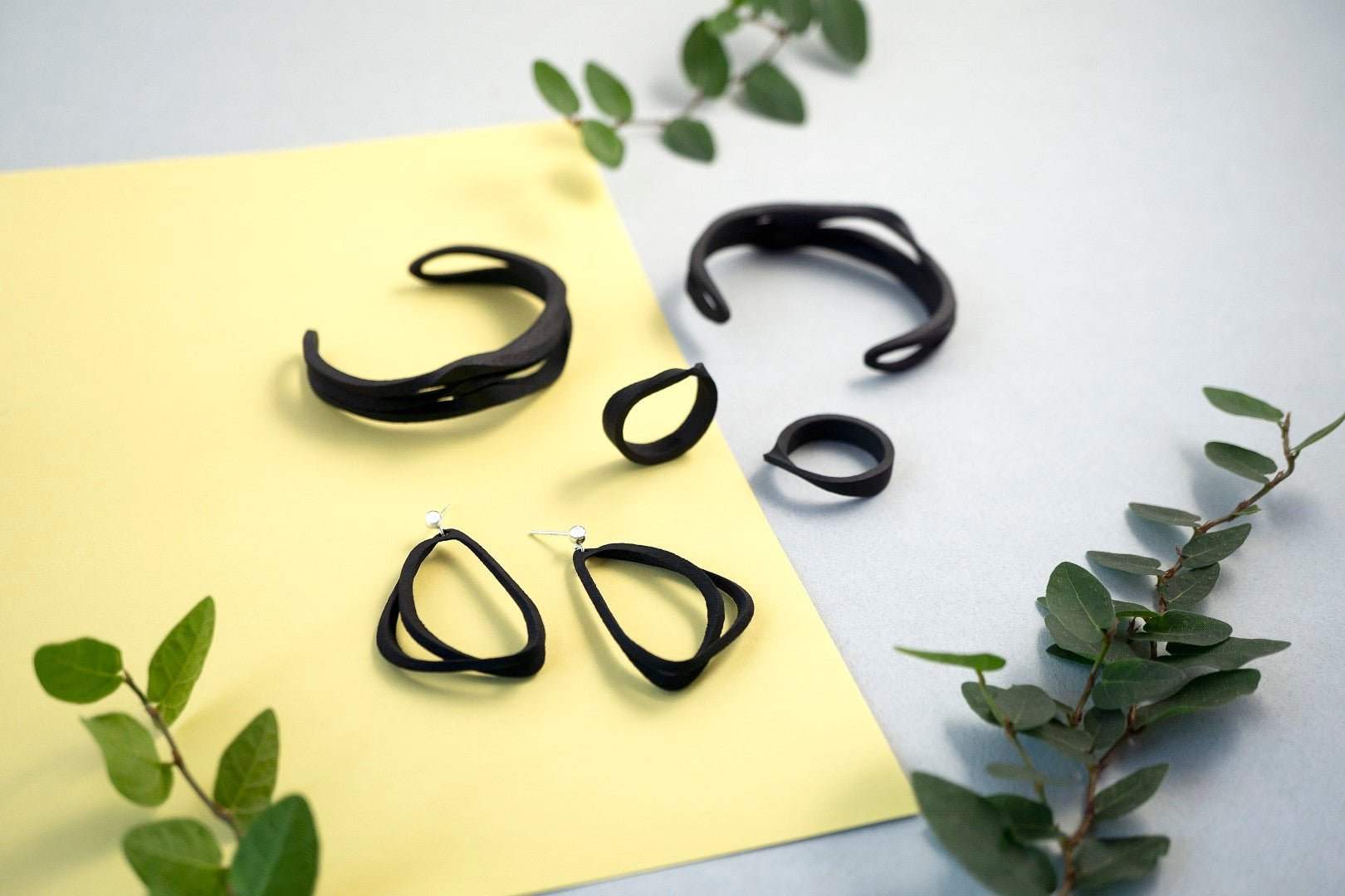 LACE Carbon Fiber Collection lightweight black carbon fiber jewelry set with earrings, rings, and cuffs on yellow and gray background with green leaves