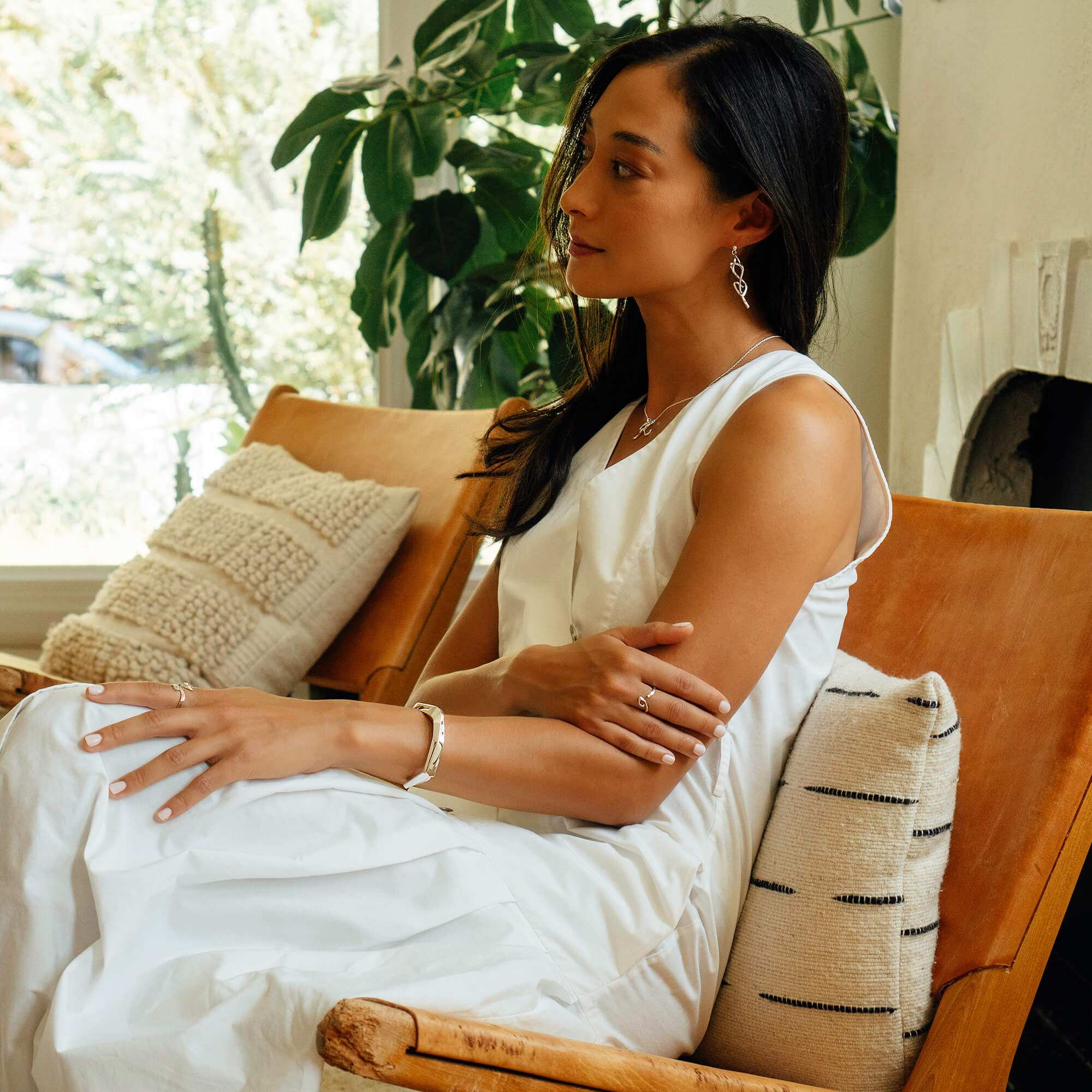 Woman wearing white dress sitting on chair showcasing Hydra Ring on finger with natural indoor background