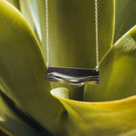 Adagio Stainless Steel Pendant with twisting curves and precision edge hanging on chain against green plant background