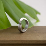 Amor Stainless Steel Ring with twisting band and precision edge detailing on wooden surface with green leaf background