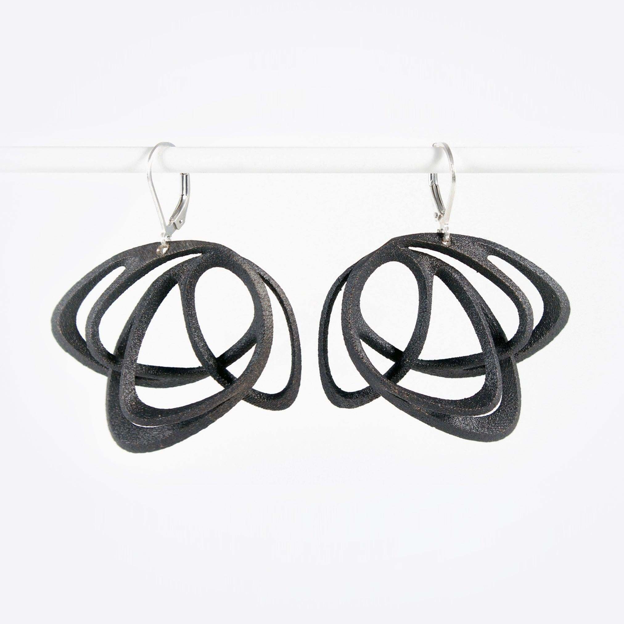 Pair of black steel Gemino earrings with interlocking elliptical petals design hanging on a white rod