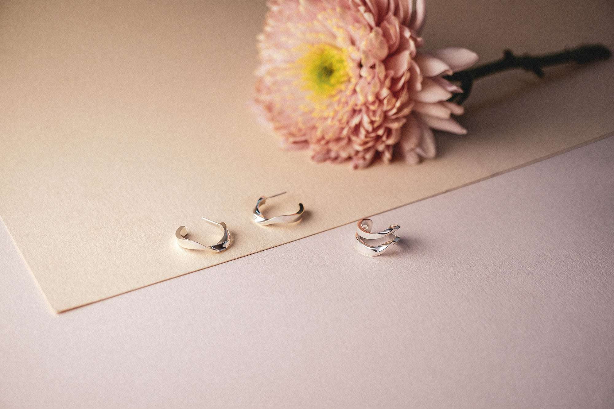 Silver Linea Earring Set with twisting bands and mini hoops on soft beige background with pink flower