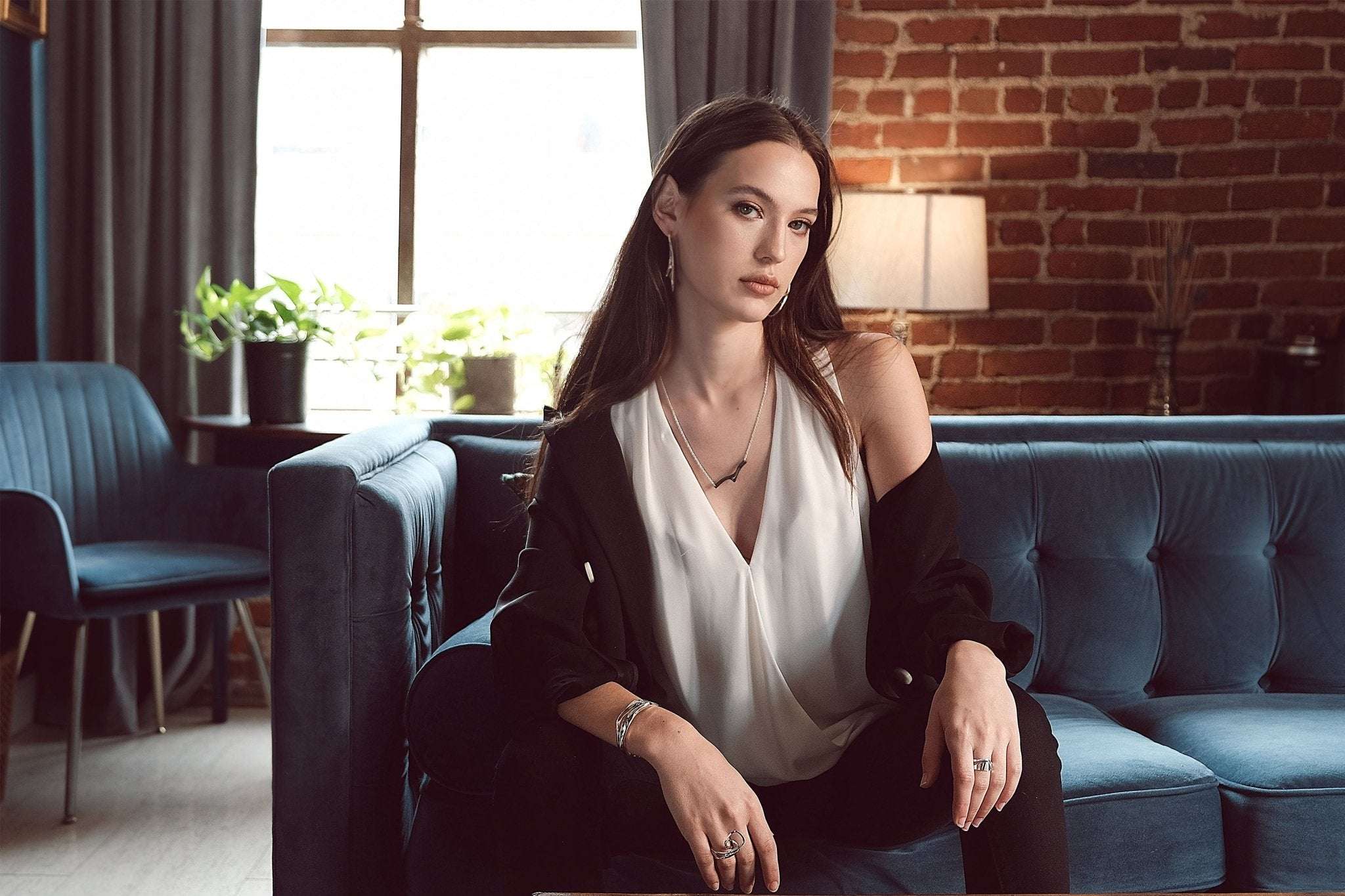 woman wearing elegant jewelry sitting on blue couch in stylish living room with brick wall European launch