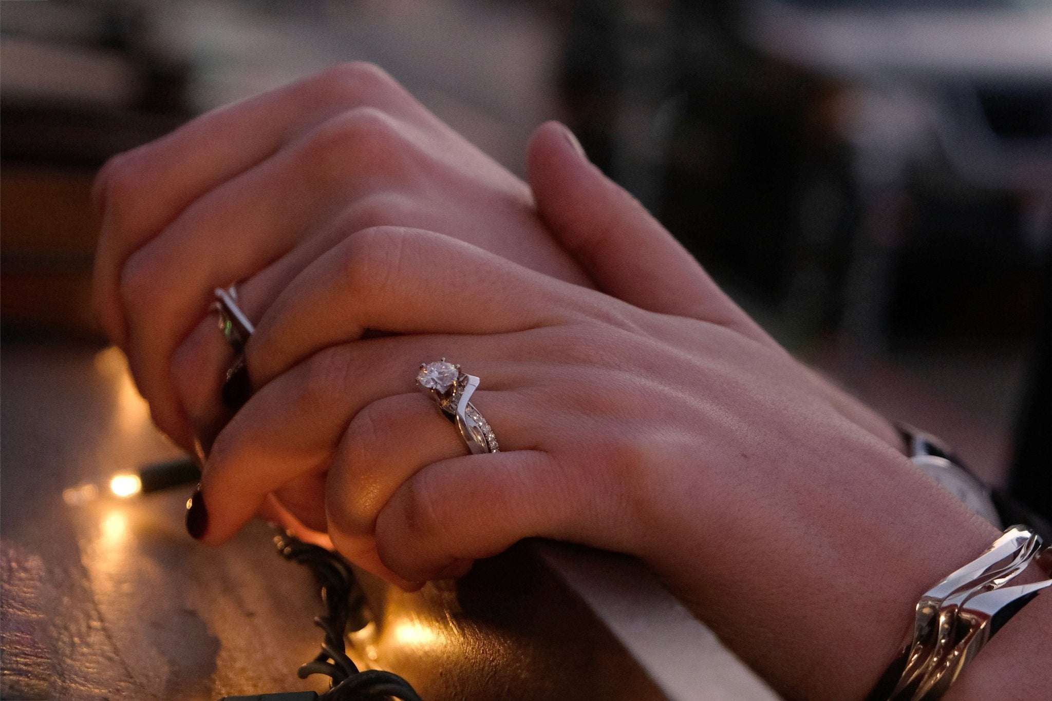 Close-up of hands with 3D printed engagement ring holding each other warmly on a wooden surface with soft lighting.