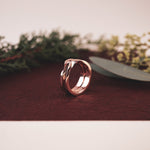 Rose gold Amor Amos Light Set stackable twisting band ring on brown surface with greenery background