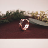 Rose gold Amor Amos Light Set stackable twisting band ring on brown surface with greenery background