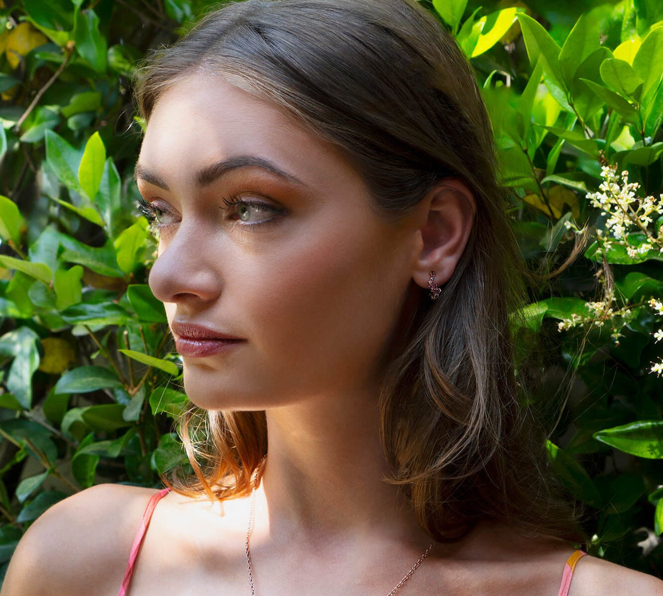 Close-up of a woman wearing delicate Leda Earrings with lilac and pink gemstones in 18K rose gold vermeil amidst green foliage.