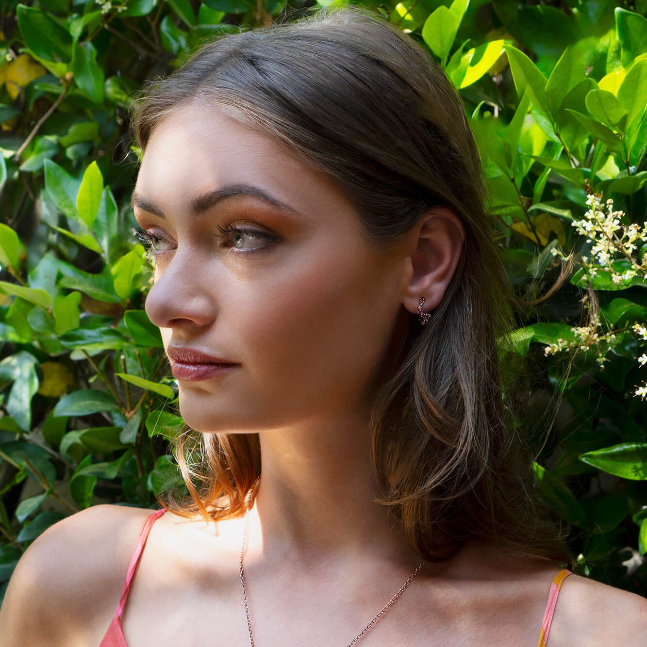 Close-up of a woman wearing delicate Leda Earrings with lilac and pink gemstones in 18K rose gold vermeil amidst green foliage.