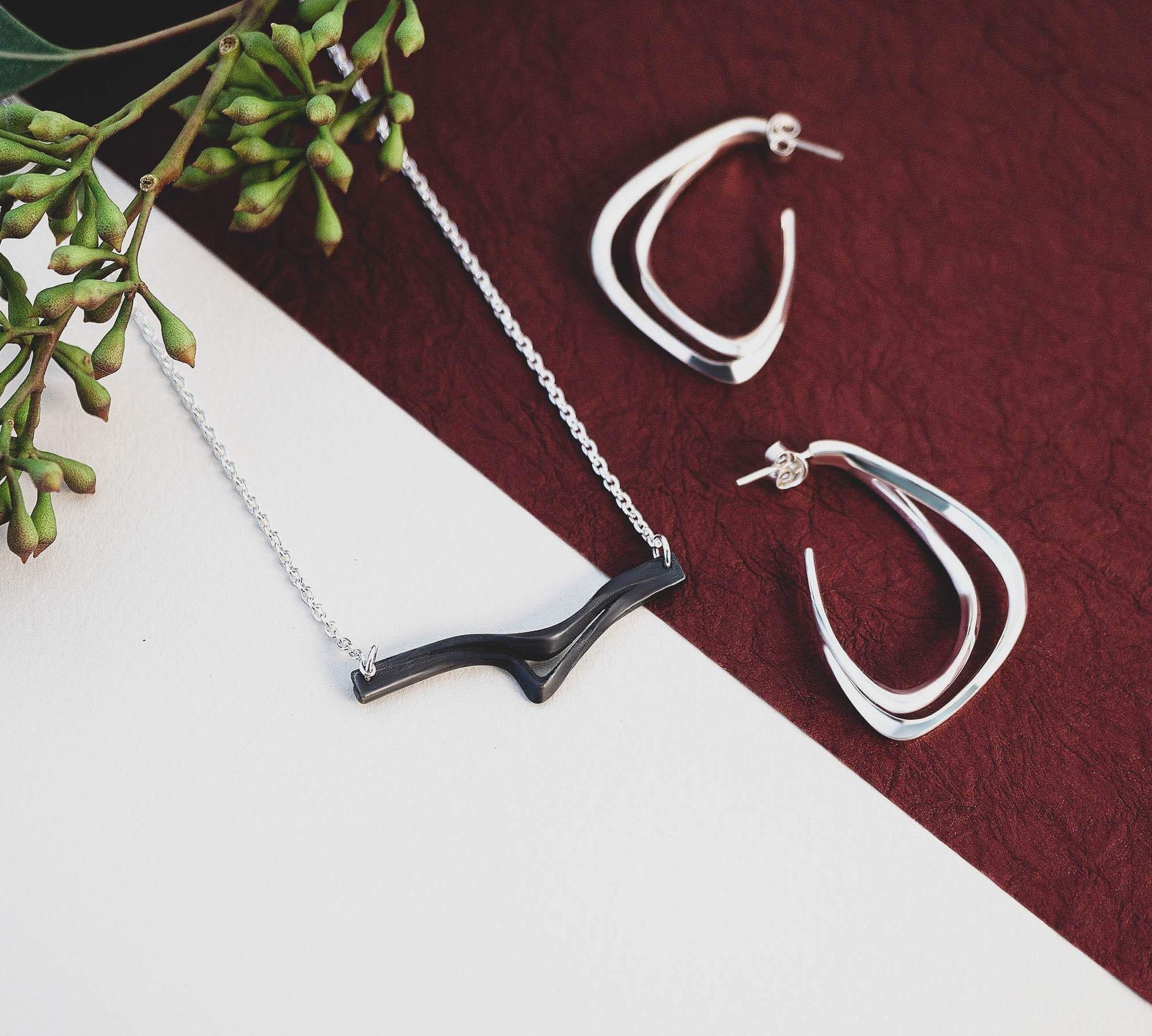 Andante Aura Set featuring steel pendant necklace and silver Aura hoop earrings on white and maroon background with green foliage